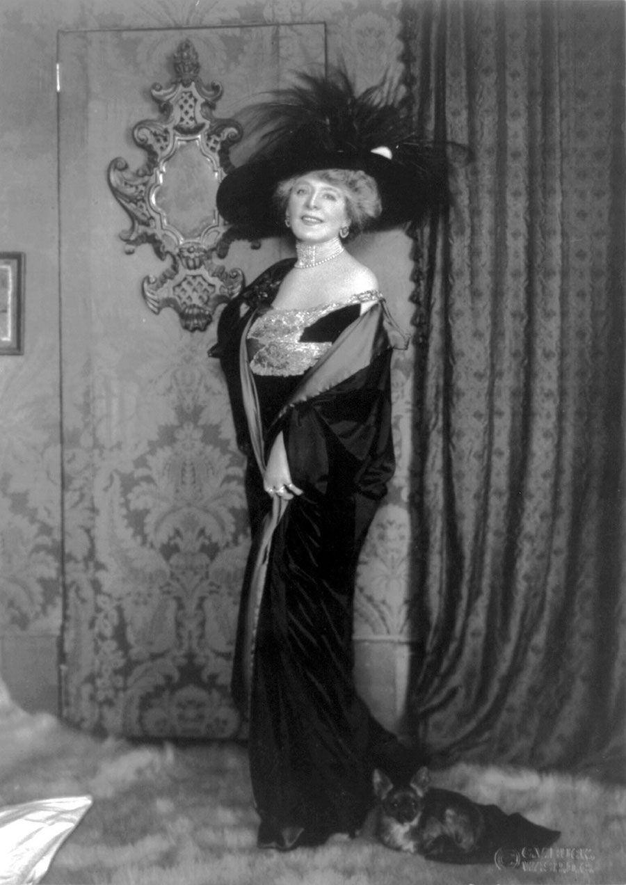 A woman in a black dress and hat holding a small dog in front of a wall with a mirror and a tapestry. A woman in a black dress and hat holding a small dog in front of a wall with a mirror and a tapestry.