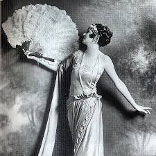 A black and white photo of a woman in a long dress and feathered headdress holding a large feather fan. A black and white photo of a woman in a long dress and feathered headdress holding a large feather fan.