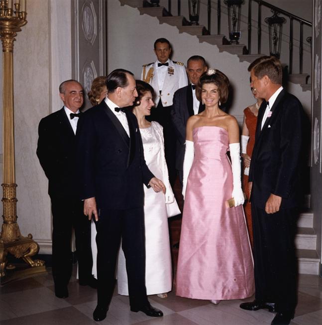 A group of people in formal attire standing in a room with a staircase in the background