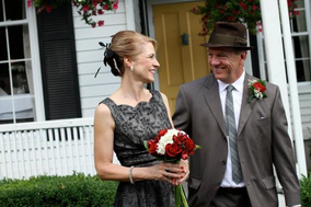 A couple dressed in formal attire standing in front of a white house with a black door and red flowers hanging from the porch. The woman is holding a red bouquet and the man has a red boutonniere. A couple dressed in formal attire standing in front of a white house with a black door and red flowers hanging from the porch. The woman is holding a red bouquet and the man has a red boutonniere.