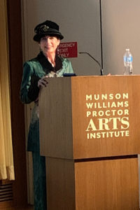 A woman standing behind a wooden podium with a microphone in a lecture hall with a red curtain and a projector screen. A woman standing behind a wooden podium with a microphone in a lecture hall with a red curtain and a projector screen.