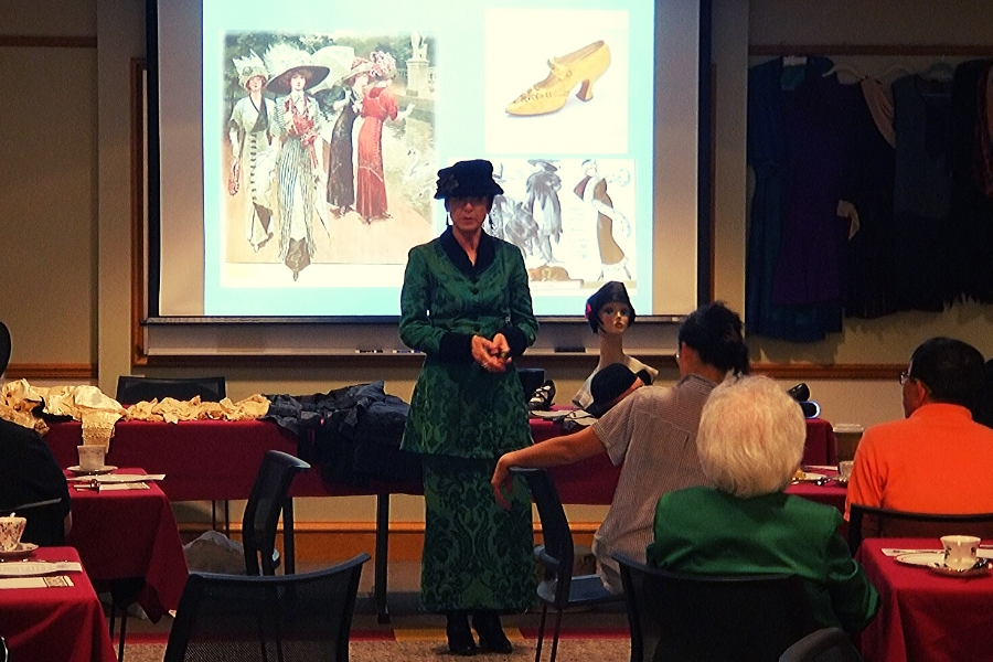 A woman in a green dress and black hat giving a presentation on historical clothing and accessories. A woman in a green dress and black hat giving a presentation on historical clothing and accessories.