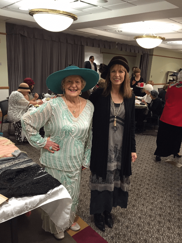 Two women wearing hats in a room with other people. The woman on the left has a green hat. The woman on the right has a black hat. They are standing in front of a table. In the background there's a stage with a man and a projector screen. Two women wearing hats in a room with other people. The woman on the left has a green hat. The woman on the right has a black hat. They are standing in front of a table. In the background there's a stage with a man and a projector screen.