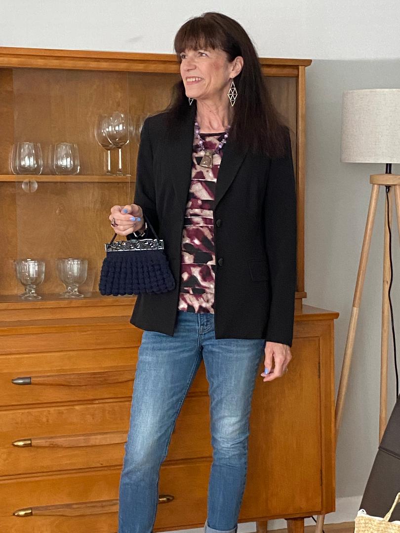 A woman in a black blazer and a pink and purple patterned top holding a small black purse with a gold chain strap. A woman in a black blazer and a pink and purple patterned top holding a small black purse with a gold chain strap.