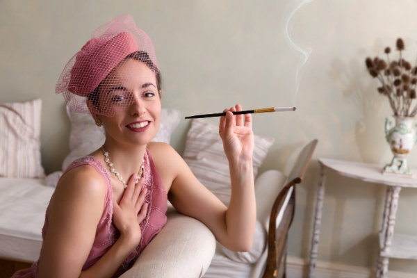 Photo realistic image of a woman in a pink dress and hat smoking a cigarette on a white bed, 1920s fashion