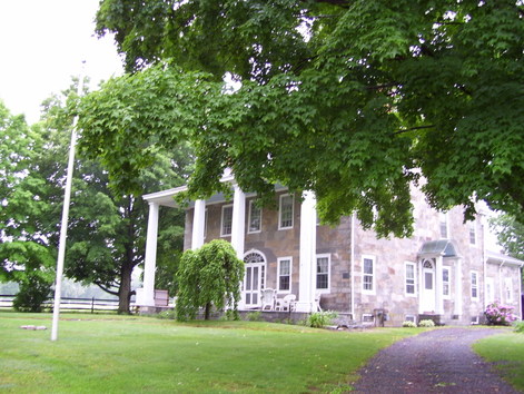 A photo of a stone house with a porch, a flagpole, and a lawn.