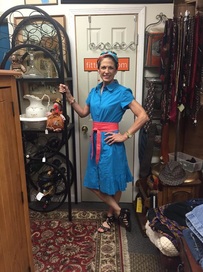 A person in a blue dress with a pink sash holding a white teapot in front of a door in an antique shop.