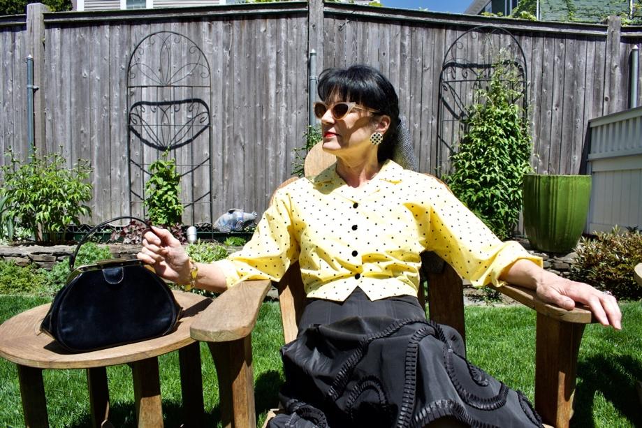 A woman in a yellow blouse and a black skirt sitting on a wooden chair in a garden A woman in a yellow blouse and a black skirt sitting on a wooden chair in a garden
